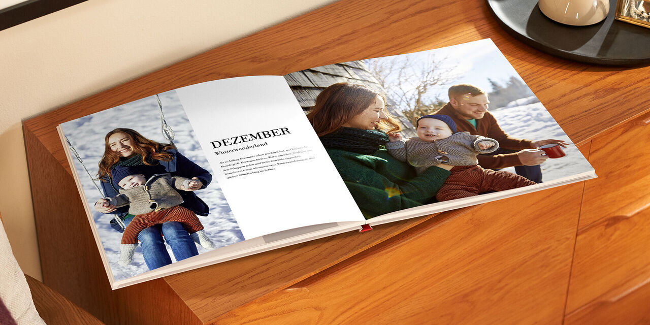 Auf einem Holzschrank liegt das aufgeklappte Fotobuch. Auf der linken Seite befinden sich ein Bild von einer Frau und einem Kind und ein Textblock mit der Überschrift «Dezember». Auf der rechten Seite ist die Familie auf einem vollflächigen Foto zu sehen.