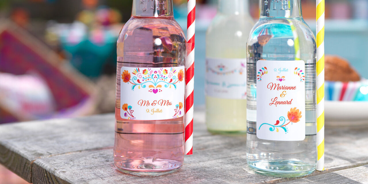 Sur une table, il y a trois bouteilles en verre avec des autocollants colorés et des pailles.