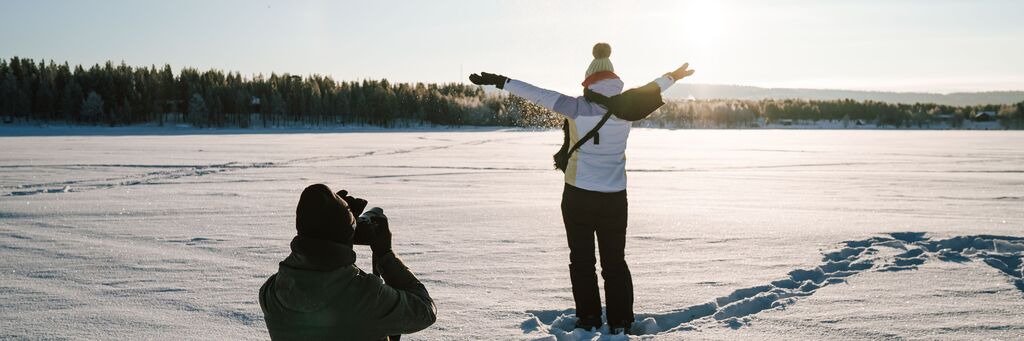 Une personne se tient sur un champ enneigé, les bras ouverts, pendant qu'une autre la photographie.