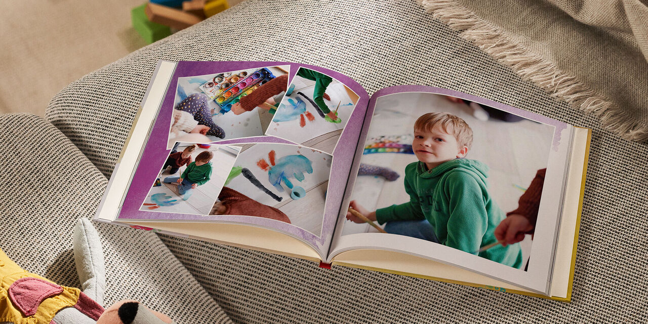 Un FOTOLIBRO CEWE aperto poggia su un divano accanto a un peluche e a una coperta. Sul lato sinistro sono visibili quattro foto leggermente sovrapposte. Tutte mostrano un bambino che dipinge un disegno con i colori di una scatola di inchiostro. La pagina di destra mostra una grande foto di un bambino che guarda nella macchina fotografica. La doppia pagina è accompagnata da un disegno ad acquerello viola.