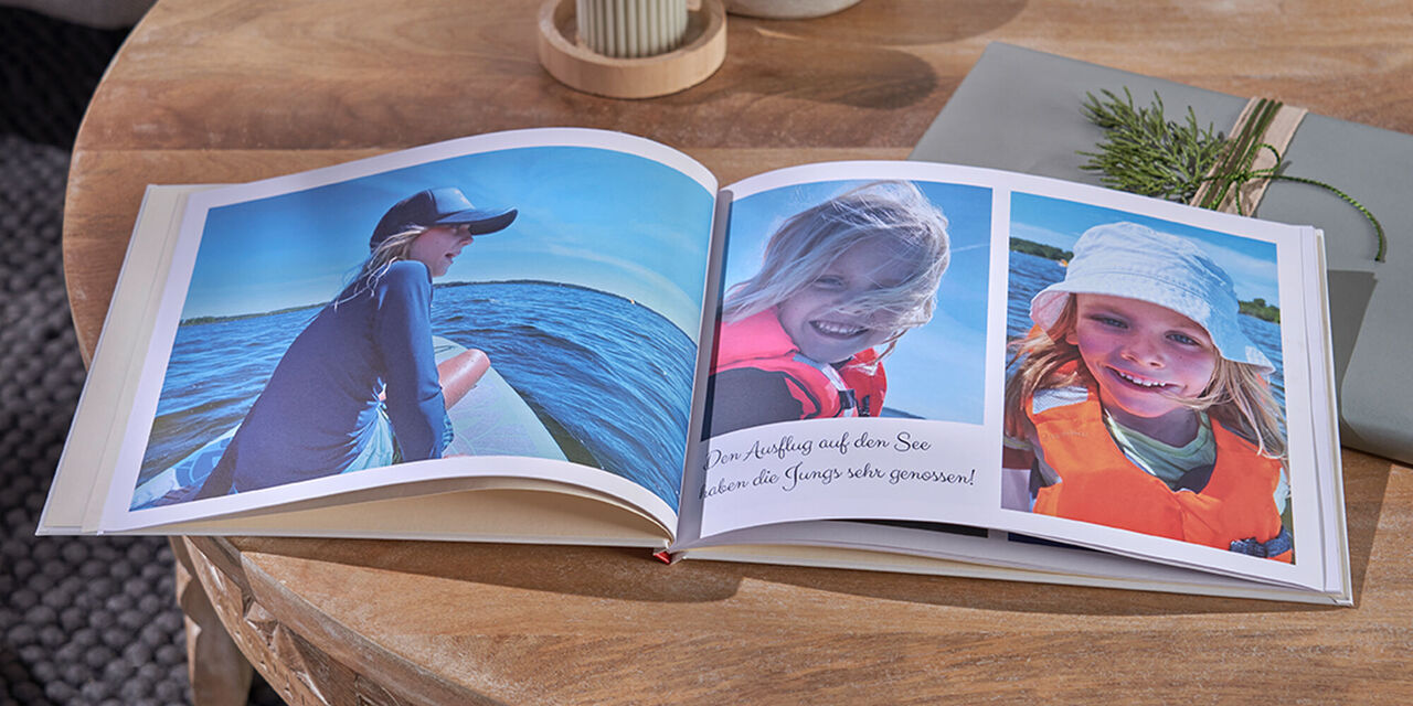 Un FOTOLIBRO CEWE aperto, due candele e un regalo giacciono su un tavolo di legno. La doppia pagina mostra le foto dei bambini su una barca.